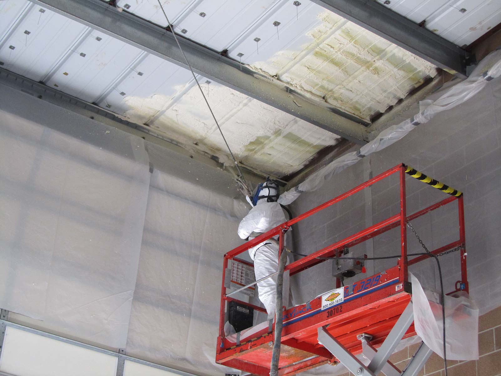 A worker on a platform spraying foam insulation into the ceiling of a metal building